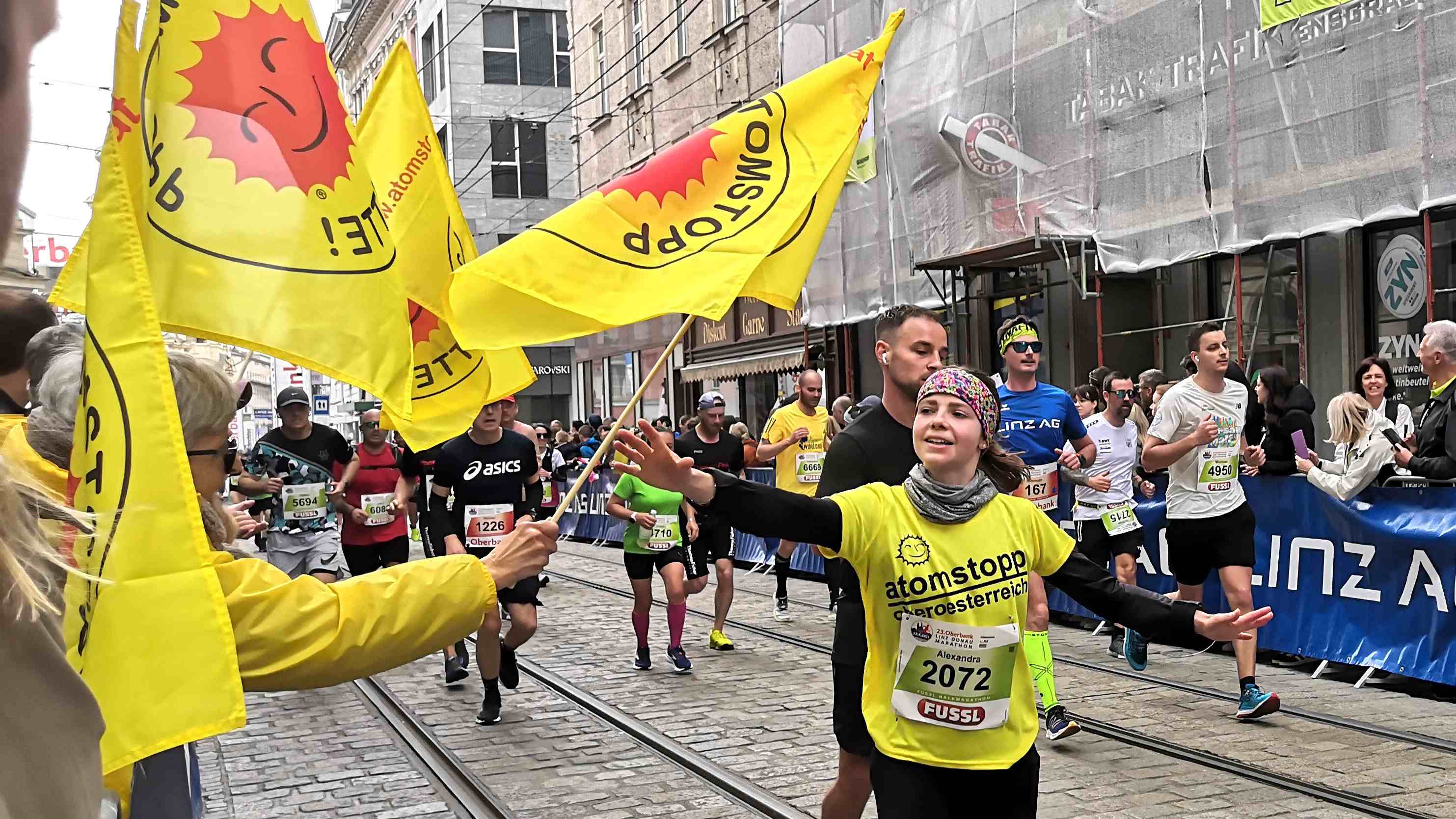 jugendliche Läuferin , etwa mit bunten Stirnband und grauem Halstuch mit gelbem Laufshirt Aufschrift atomstopp oberösterreich übernimmt Fahne www.atomstopp.at. Auf der Seite Publikum und die Hände von atomstopp-Helfer_innen mit weiteren Fahnen.