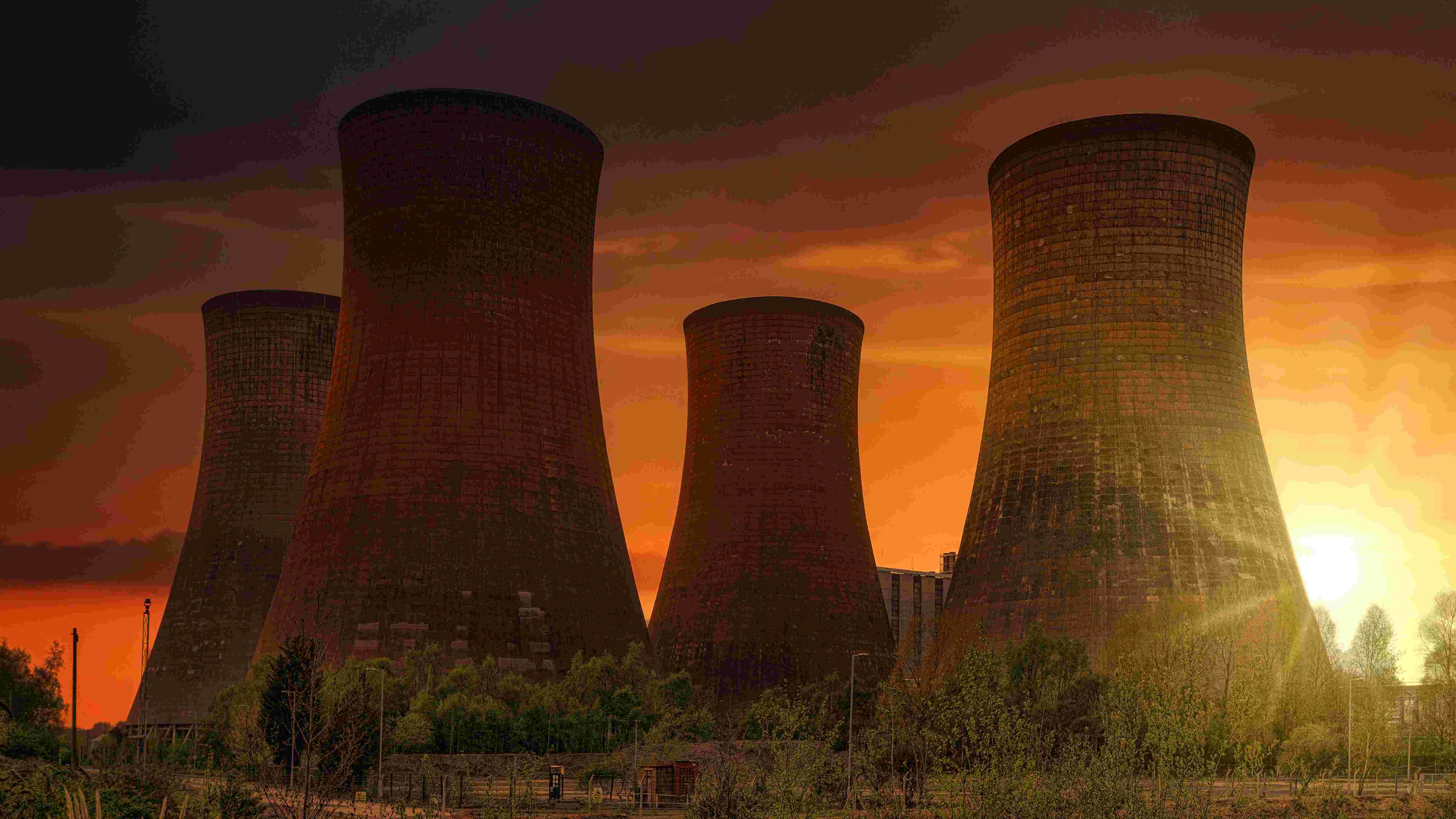 Vier große Kühltürme eines AKW ragen vor einem orange-rot leuchtenden Sonnenuntergang in den Himmel. Sie dampfen nicht, das AKW ist abgeschaltet. Im Vordergrund befinden sich Bäume und Vegetation, während die tief stehende Sonne rechts neben den Türmen sichtbar ist.