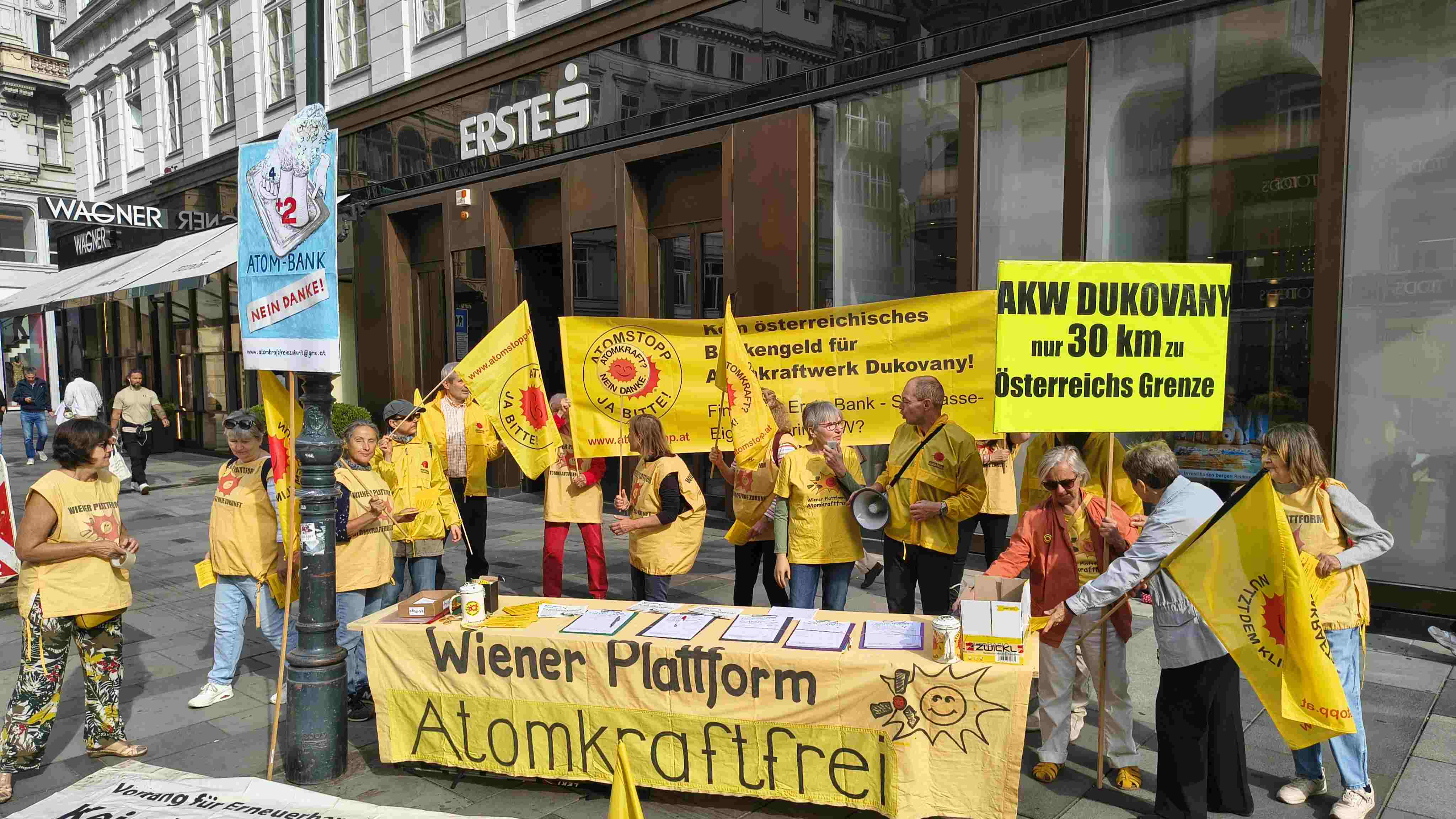 Demonstrant_innen mit Fahnen und einem großen gelben Banner vor der Erste Bank - Sparkasse am Wiener Graben. Das Banner ist 3 mal 1 Meter groß, gelb mit schwarzem Text "Kein österreichisches Bankengeld für Atomkraftwerk in Dukovany. Finanziert Erste Bank -Sparkasse-Eigentümerin AKW?" und hat links die rote Anti-Atom-Sonne abgebildet.