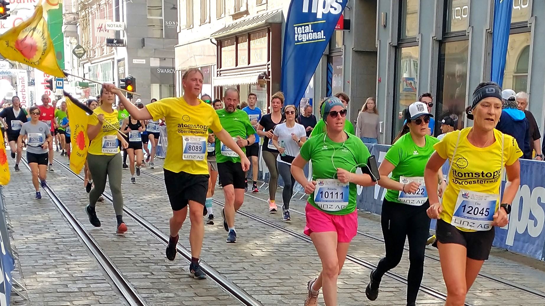 mehrere Läufer und Läuferinnen mit gelben Laufshirts und Fahnen beim Linz Marathon