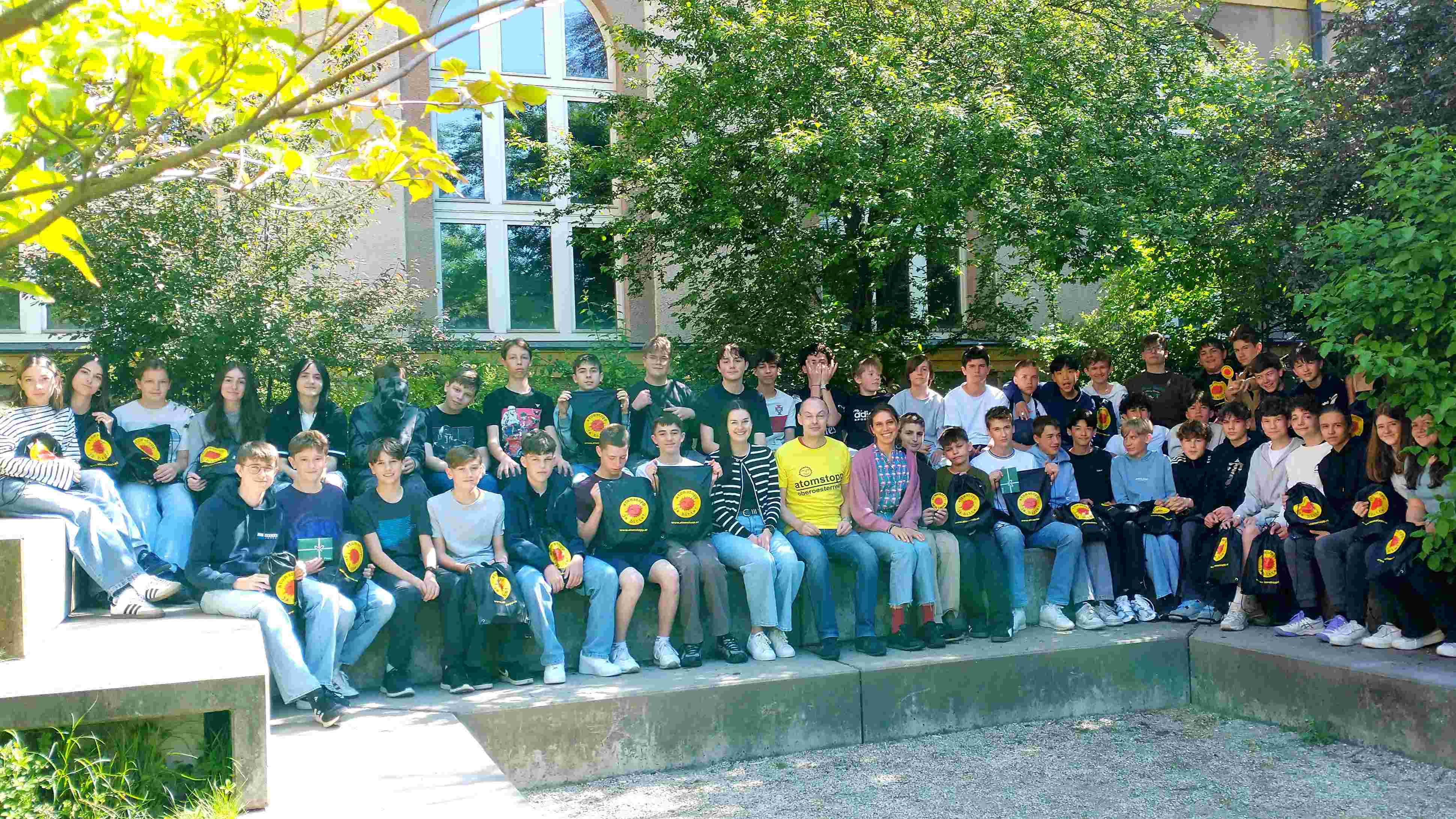 Etwa 30 Schüler_innen mit 2 Lehrerinnen und atomstopp Geschäftsführer Stoiber sitzend unter Bäumen mit Goodiebags mit dem Logo atomstopp - ja bitte!