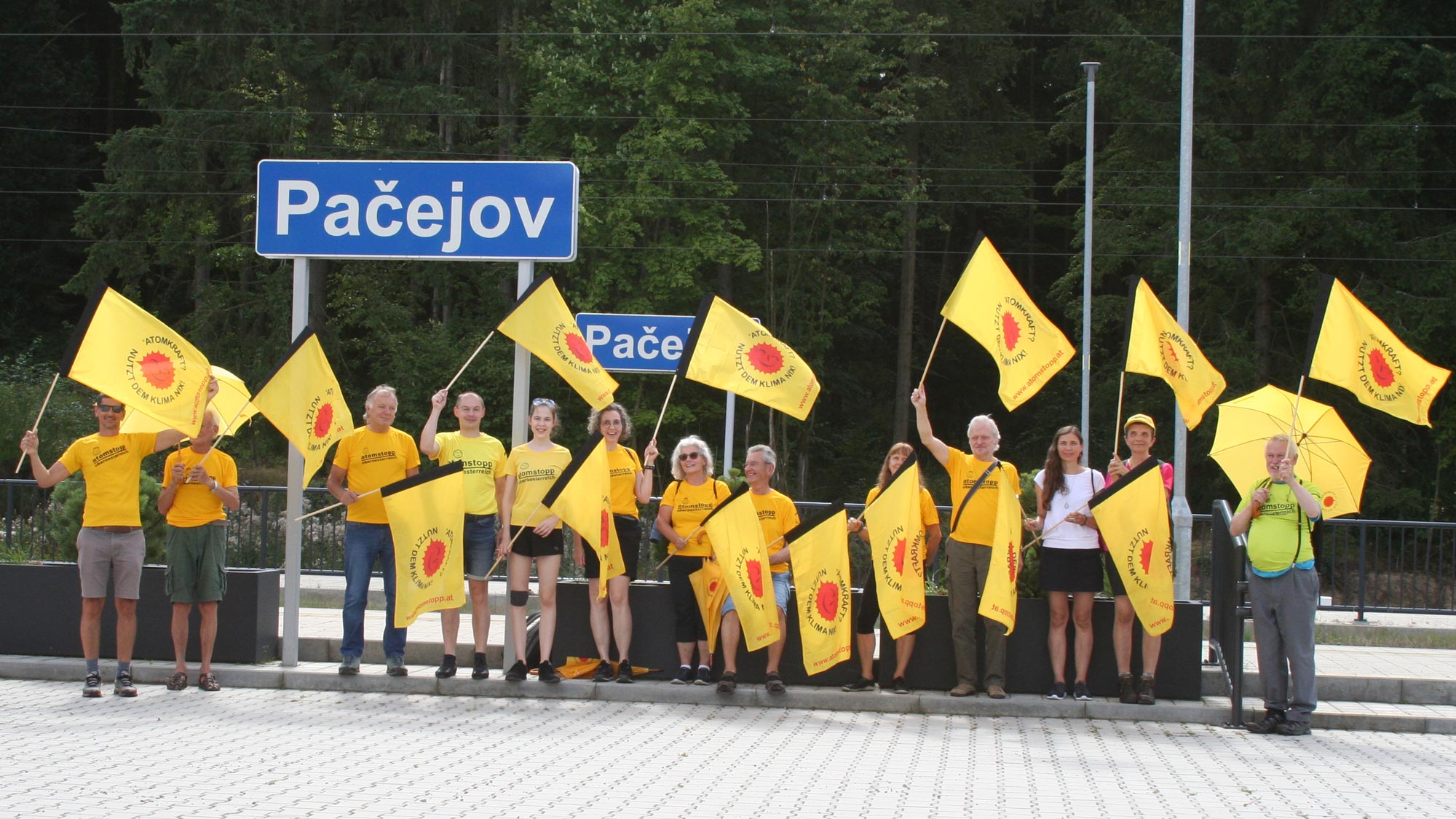 Unterstützer_innen in sonnengelben atomstopp-Shirts und mit wehenden Fahnen bei der Protest-Wanderung, vor dem Stationsschild "Pacejov"