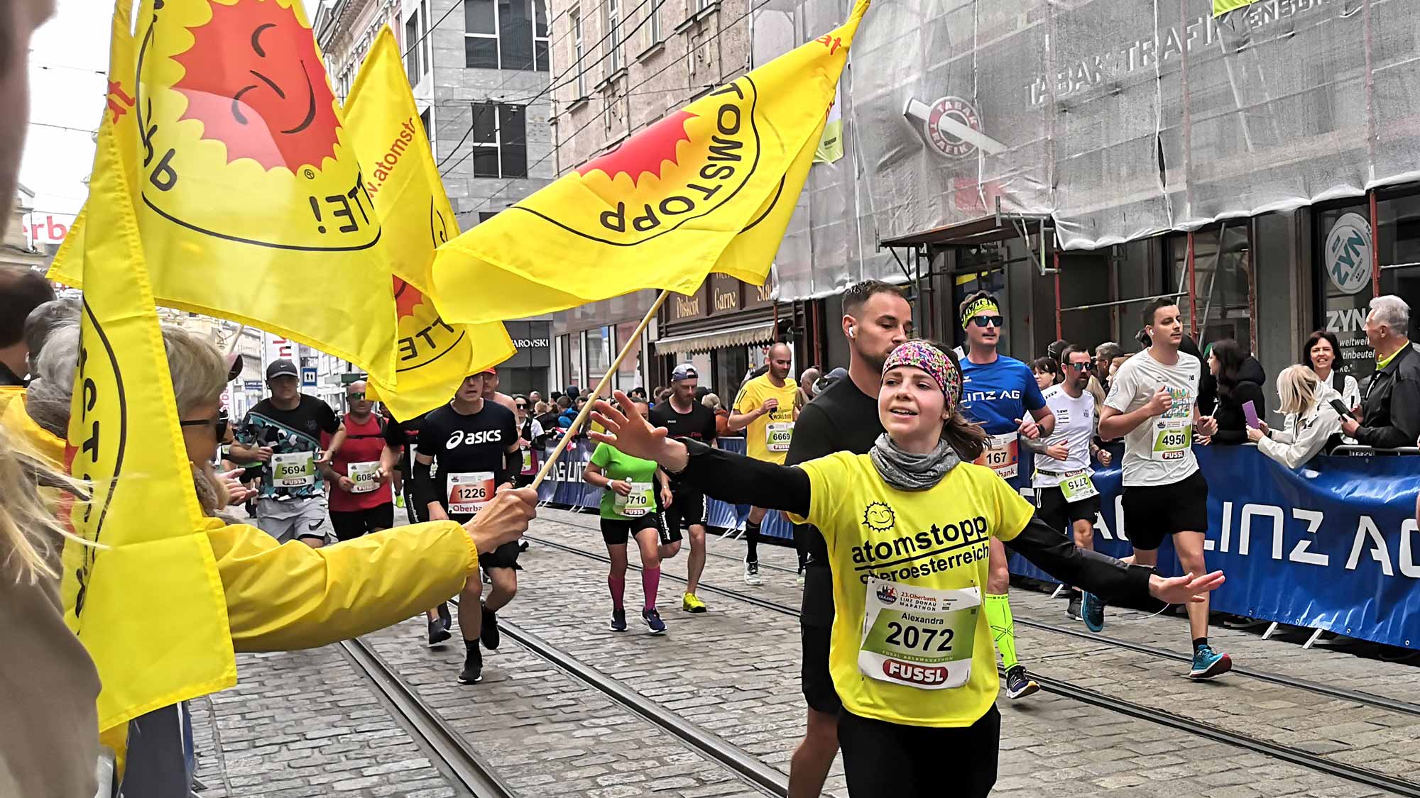 Eine Läuferin in gelbem atomstopp-Shirt greift nach der gelben Fahne mit atomstopp-Logo (rote, lachende Sonne mit Schriftzug "Atomstopp ja bitte!" für den Zieleinlauf beim Linz Marathon